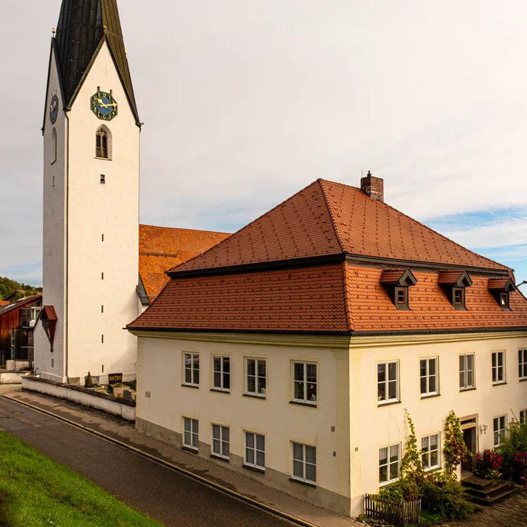 Kirche St. Martin in Martinszell – historische Pfarrkirche mit hohem, spitzem Kirchturm und rotem Ziegeldach, umgeben von Wohnhäusern am Hang bei Waltenhofen.