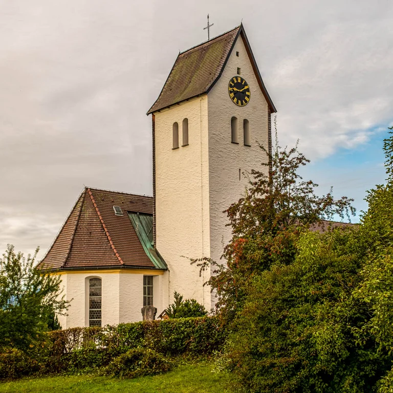 Kirche St. Andreas in Memhölz – kleine, historische Dorfkirche mit hellem Putz, rotem Ziegeldach und markantem Kirchturm mit Uhr, umgeben von grüner Landschaft.